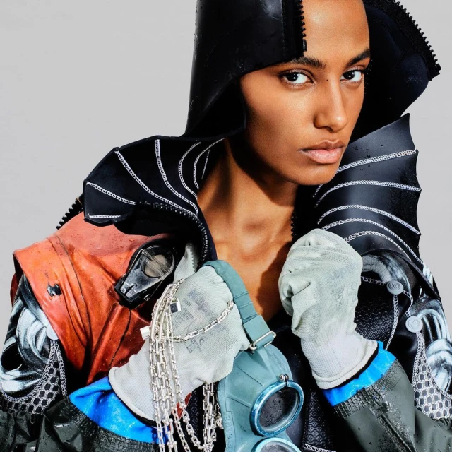 Portrait of an adult female fashion model wearing a black hooded jacket and holding a gas mask, standing in front of a solid grey background.