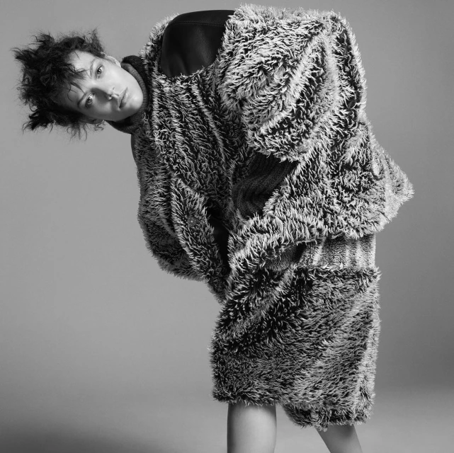 Monochrome portrait of a woman wearing a fuzzy coat and skirt, leaning forward and looking at the camera, with a grey studio background.