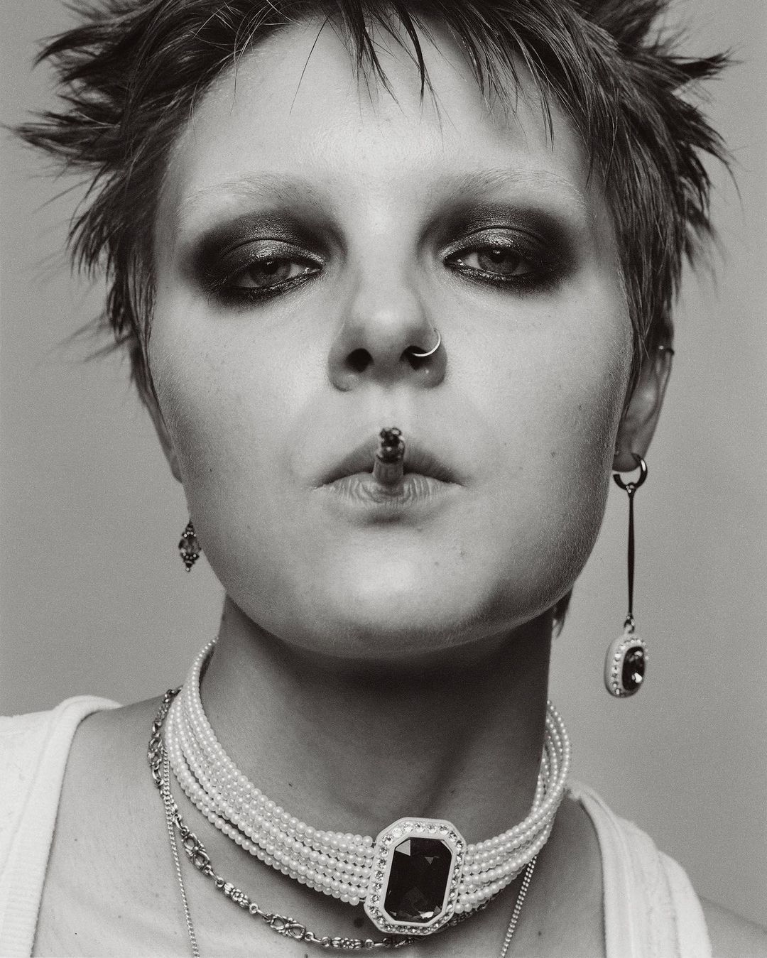 Striking black and white portrait of a woman smoking, adorned with pearl and gemstone jewelry, and bold, dark eyeshadow.