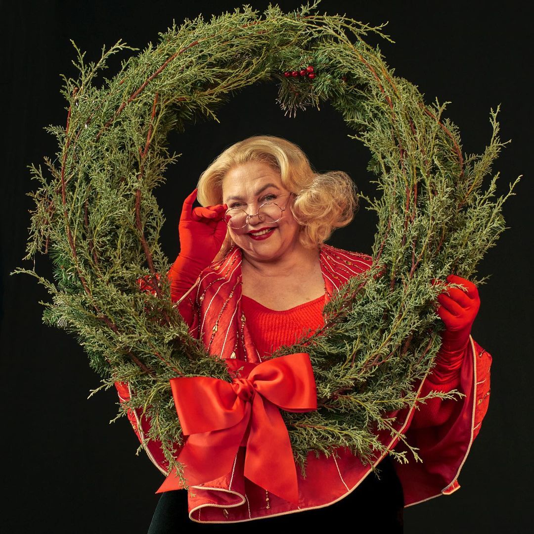 Mature woman smiles while holding a circular juniper wreath decorated with a red bow and berries against a dark background.
