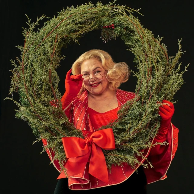 Mature woman smiles while holding a circular juniper wreath decorated with a red bow and berries against a dark background.