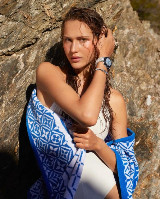 Woman poses near a rock wearing a bathing suit and a watch with a blue and white towel on a sunny day.