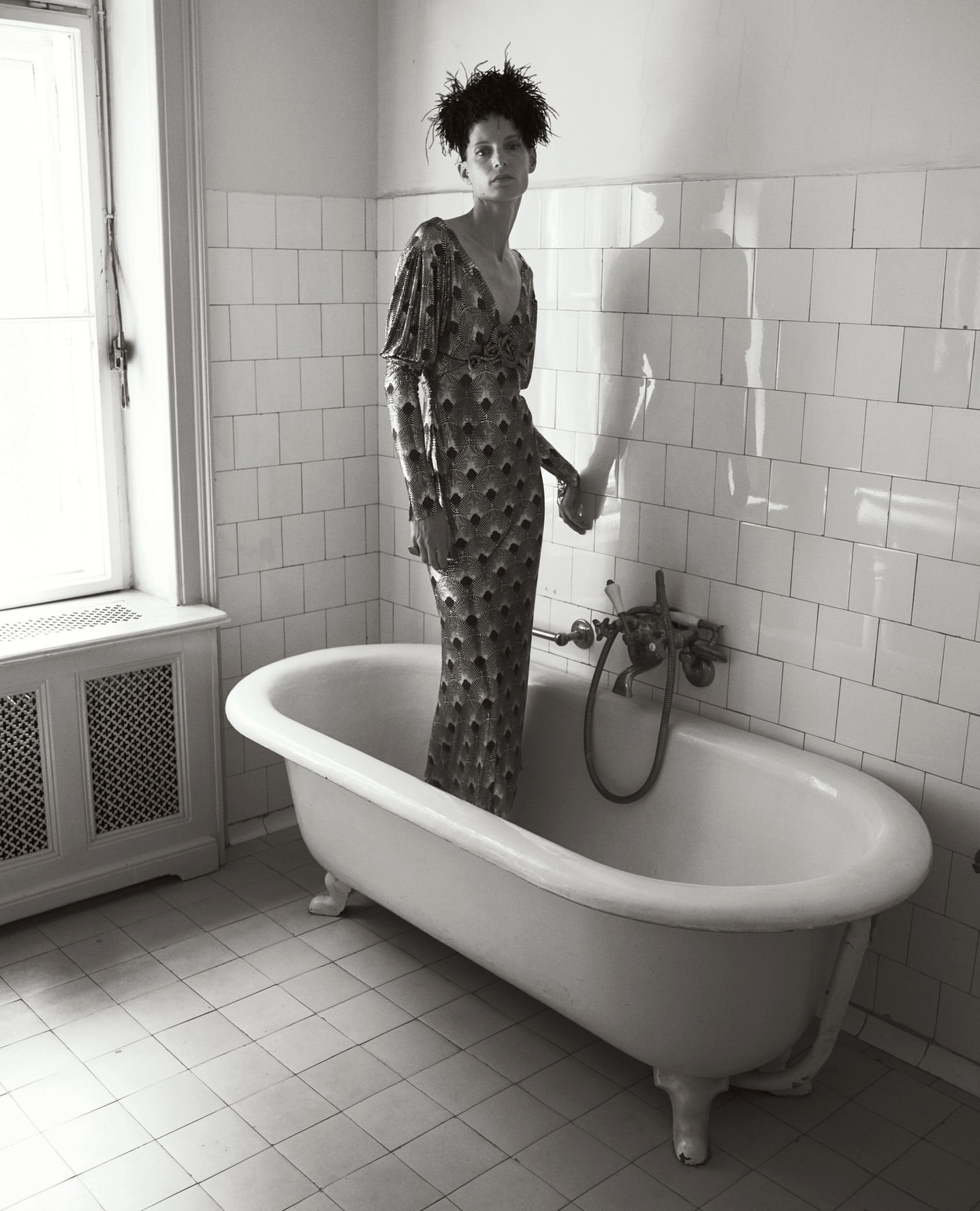 A woman in a dress stands in a bathtub in a tiled bathroom. The black and white photo shows classic bathroom fixtures and minimalist style.
