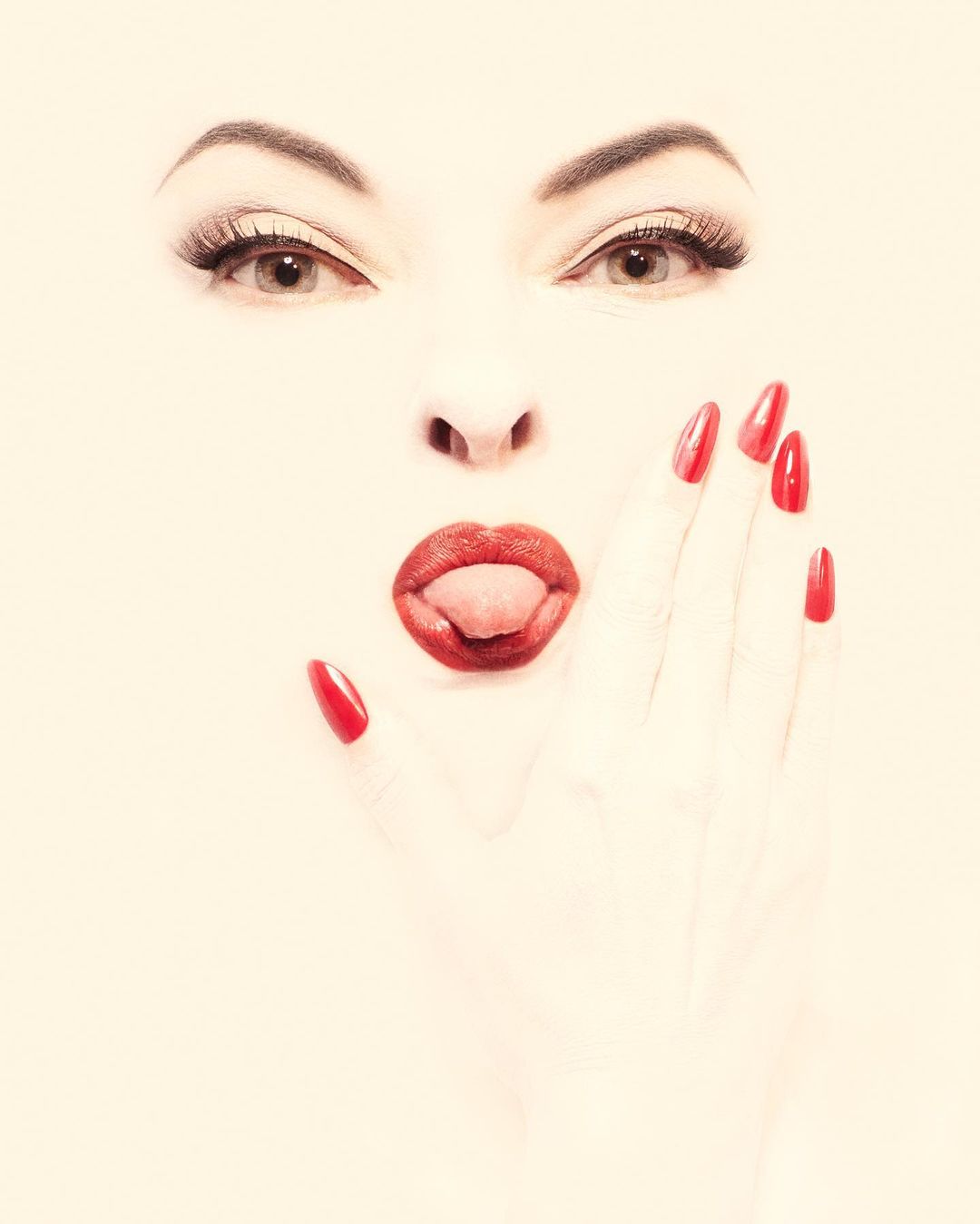 Close-up portrait of a woman with bright red lipstick and long red stiletto fingernails holding her hand to her cheek.