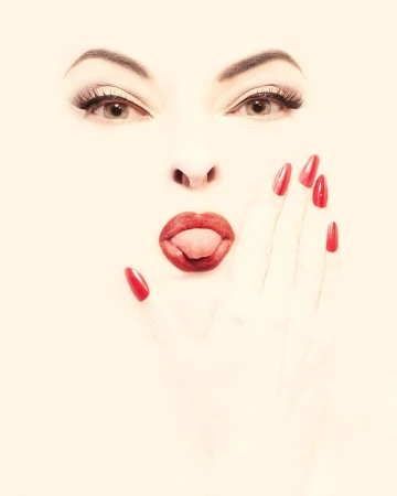 Close-up portrait of a woman with bright red lipstick and long red stiletto fingernails holding her hand to her cheek.