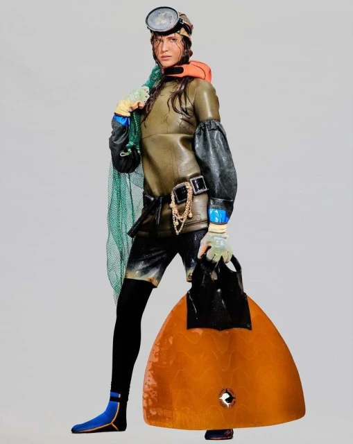 Adult female model in a scuba suit and dive gear with a fishing net and a large diving fin poses in a full shot against a grey backdrop.