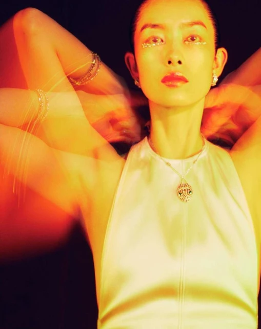 An Asian woman with glitter makeup poses with hands behind her head, wearing a white top and jewelry, against a dark background.