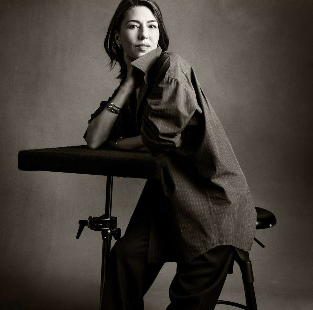 Elegant portrait of a woman in an oversized pinstripe shirt, resting her head on her hand while seated in front of a grey backdrop.
