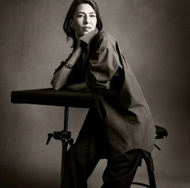 Elegant portrait of a woman in an oversized pinstripe shirt, resting her head on her hand while seated in front of a grey backdrop.