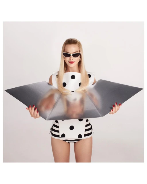 Blonde woman in retro swimwear holds a reflective screen in a white photo studio, wearing sunglasses and red lipstick.