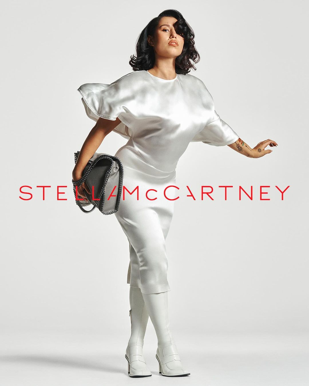 A stylish adult woman with dark hair poses confidently in a studio wearing white apparel, knee-high boots, and holding a Stella McCartney handbag.