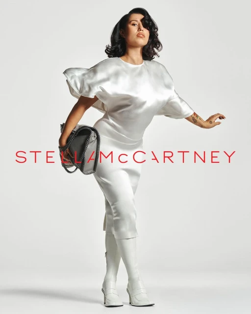 A stylish adult woman with dark hair poses confidently in a studio wearing white apparel, knee-high boots, and holding a Stella McCartney handbag.