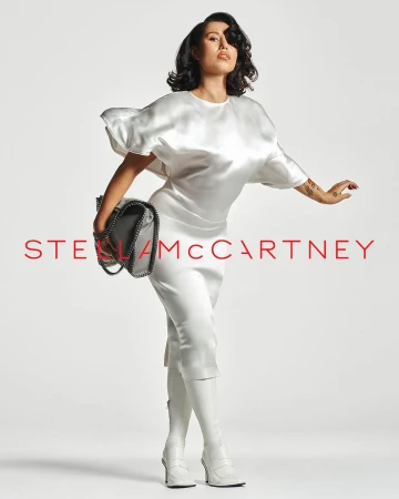 A stylish adult woman with dark hair poses confidently in a studio wearing white apparel, knee-high boots, and holding a Stella McCartney handbag.