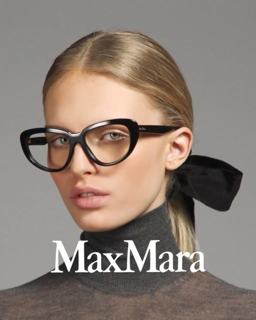 A close-up studio shot of a blonde woman wearing Max Mara glasses, a grey turtleneck, and a black hair ribbon against a grey backdrop.