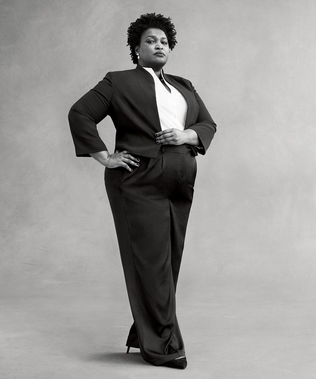 Full body portrait of a Black woman in a dark pantsuit with hands on her hips, posing in front of a plain gray background.