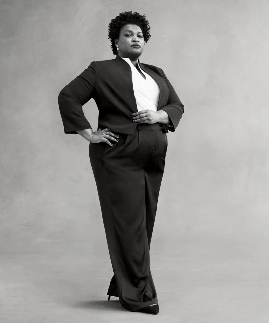 Full body portrait of a Black woman in a dark pantsuit with hands on her hips, posing in front of a plain gray background.