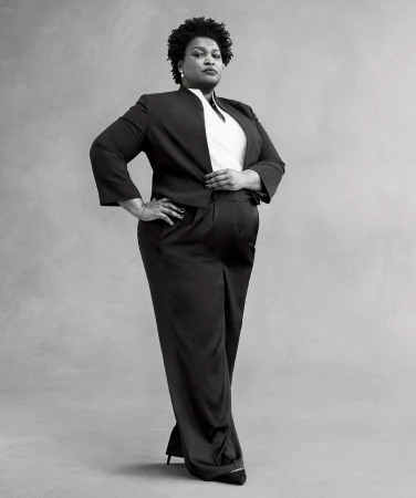 Full body portrait of a Black woman in a dark pantsuit with hands on her hips, posing in front of a plain gray background.