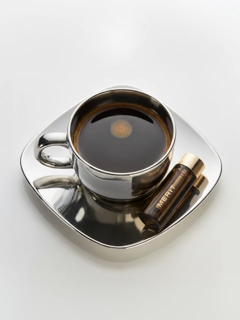 A silver cup of black coffee sits on a matching saucer, next to a Merit brand cosmetic tube on a plain white surface in studio lighting.