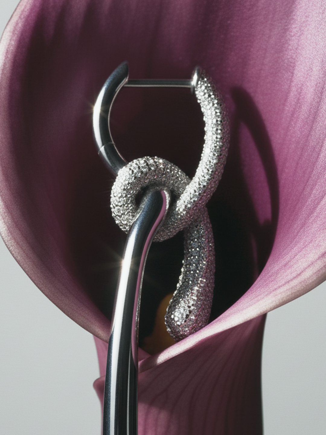 Close-up of a metal hoop earring featuring a textured, shimmering section, intertwined with a smooth metal accent inside a purple calla lily.