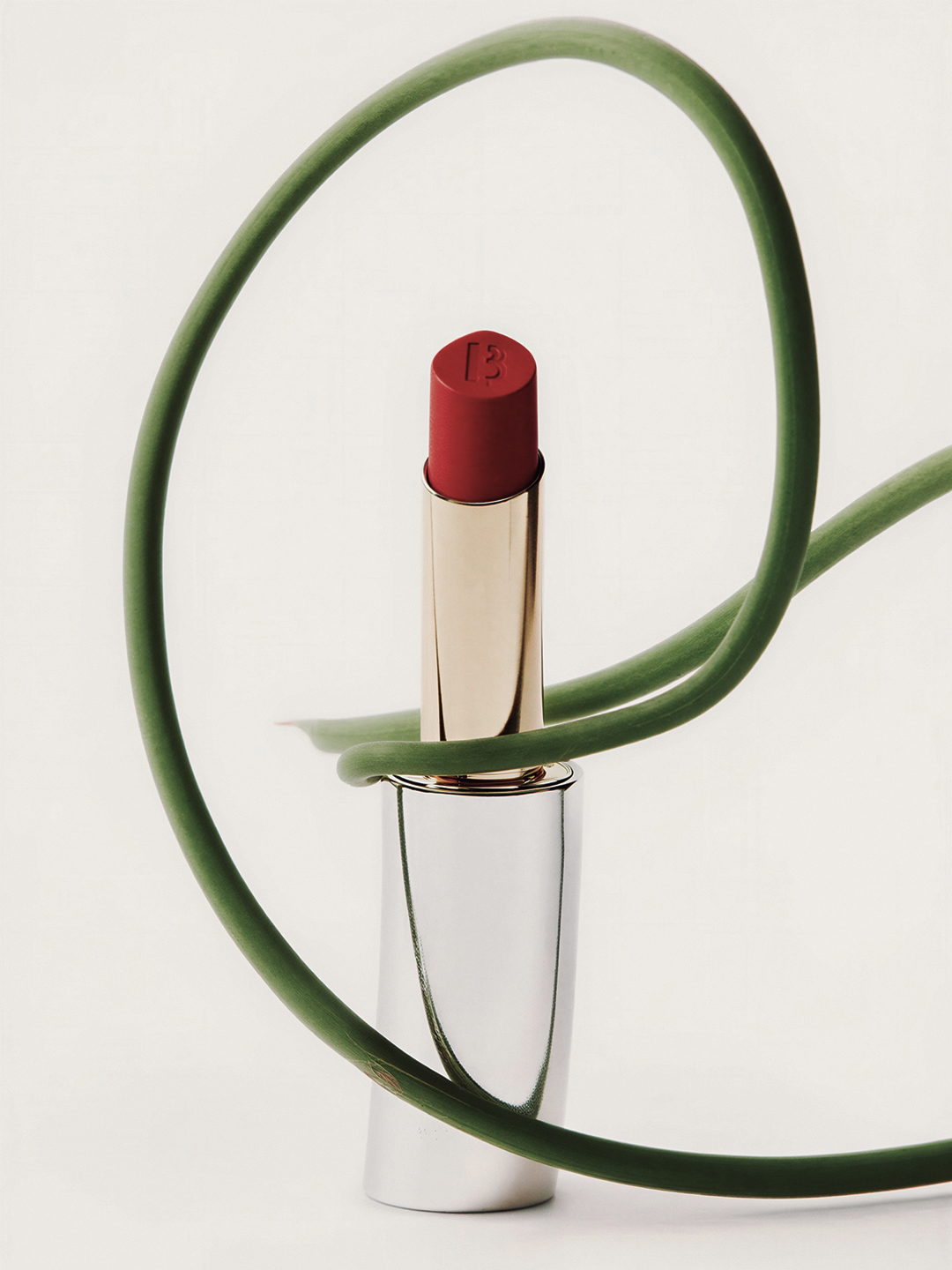 Studio shot of a red lipstick with gold and silver tube, encircled by a green plant stem, against a white background.