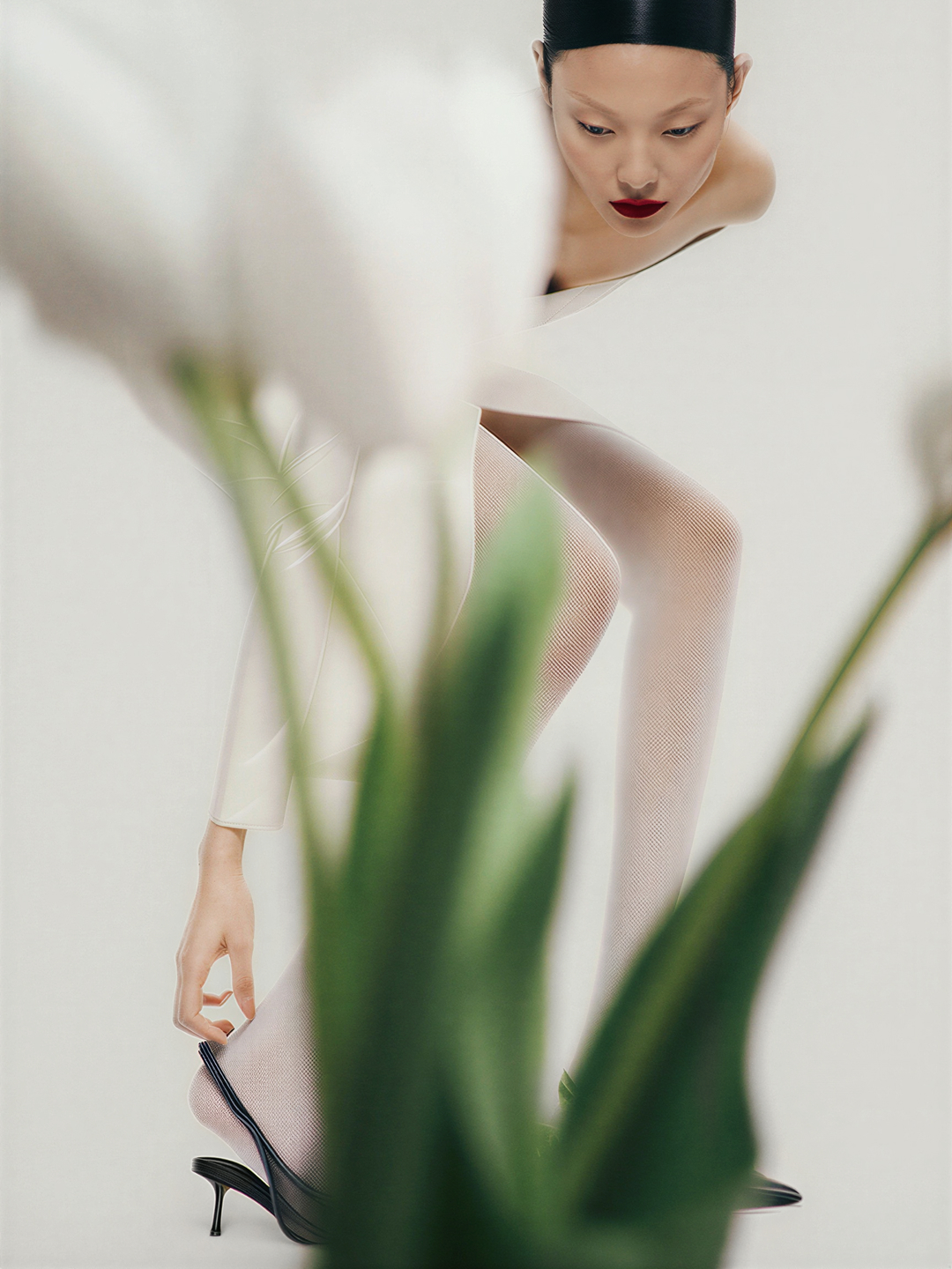 Woman adjusting black high heel shoe with floral foreground in a fashion shot with bright studio lighting and a clean aesthetic.