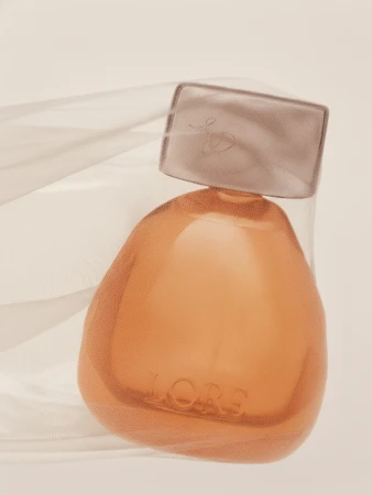 Amber glass perfume bottle embossed with LORE wrapped in sheer white fabric against a warm neutral background.