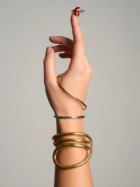 Hand with multiple gold and silver bracelets has a ladybug on the tip of a finger on a neutral grey background in studio lighting.