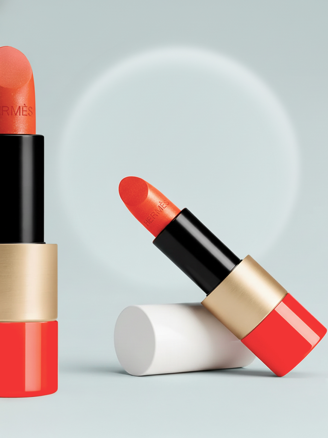 Close-up of orange Hermes lipstick with black and gold case with lid on a pale blue surface with a circular light.