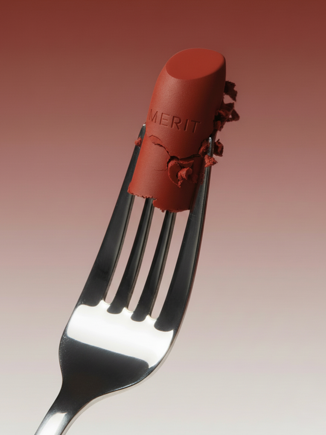 Close-up of an unwrapped, partially broken red lipstick with the word Merit on it, speared on a silver fork against a gradient background.