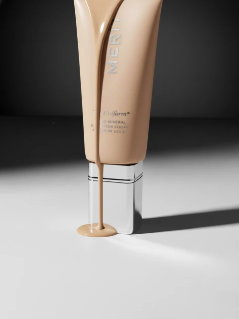 Merit makeup tube with light-brown liquid foundation, on a white surface against a gray background. Beauty product close-up.