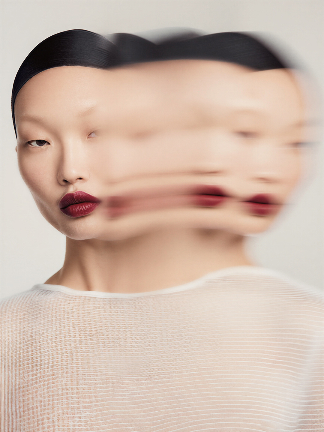 Asian woman with sleek dark hair and dark red lipstick in a fashion beauty shot with a blurred effect against a plain background.