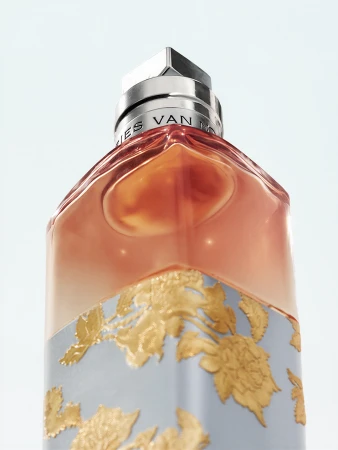 Close-up studio shot of a stylish glass perfume bottle with gold floral designs and chrome accents against a clean, light blue background.