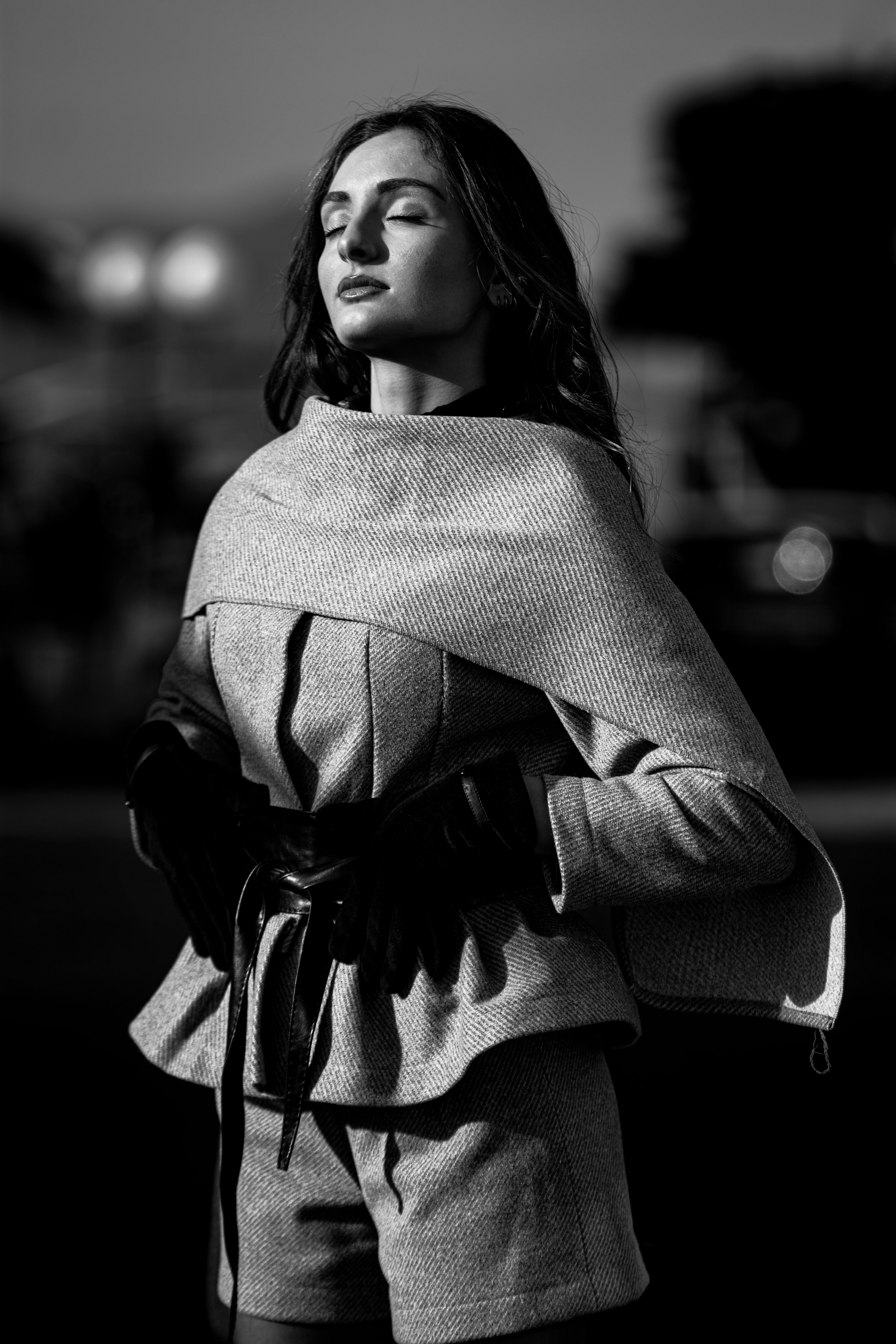 Black and white portrait of a female fashion model wearing a tweed jacket with short shorts, eyes closed, standing outside.