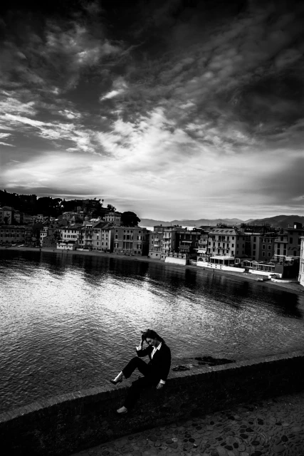 Adult female in dark suit sitting on stone wall by the water, buildings across the water, cloudy sky, black and white photograph.