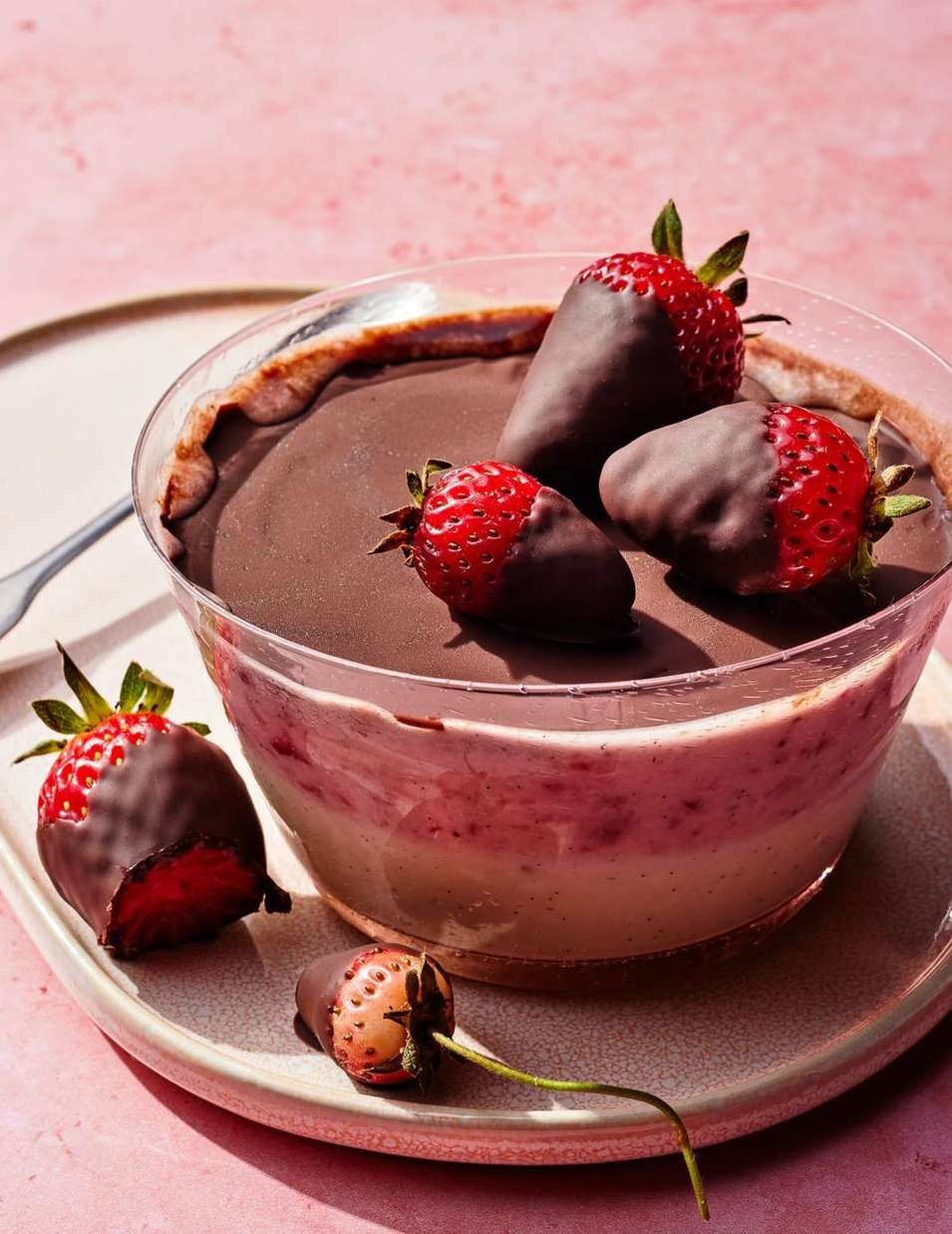 Close-up of chocolate-covered strawberries on a dessert in a clear bowl, atop a plate against a pink background. Food photography.