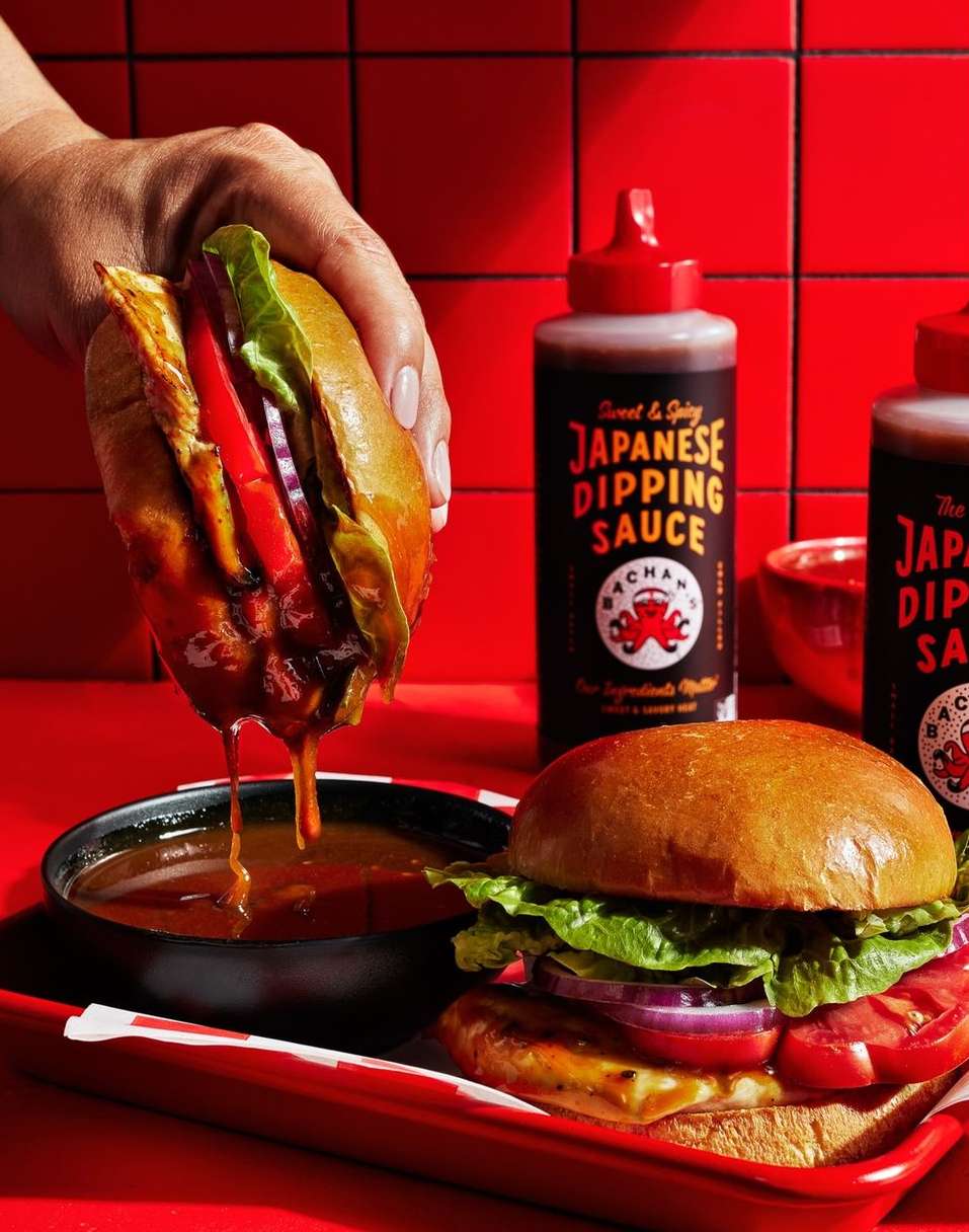 Two burgers with visible toppings next to Japanese dipping sauce, presented on a red tray against a red tiled wall backdrop.