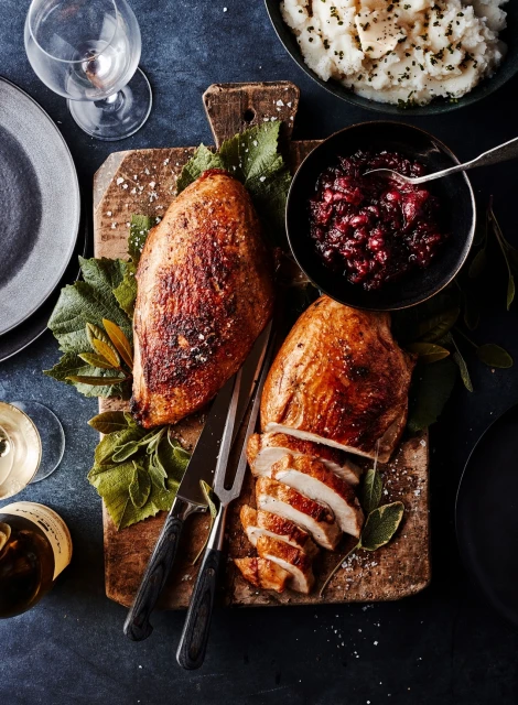 Overhead shot of sliced turkey breasts, cranberry sauce, and mashed potatoes on a wooden board with plates and wine glass.