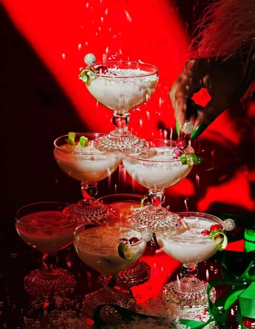 Champagne coupe tower with snow-like sprinkles and lime twists, set against a bold red backdrop, exudes celebratory elegance.