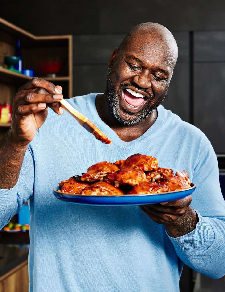 Shaquille O'Neal smiles widely holding plate of chicken wings with BBQ sauce, in kitchen, wearing blue top, indoor setting, close-up.