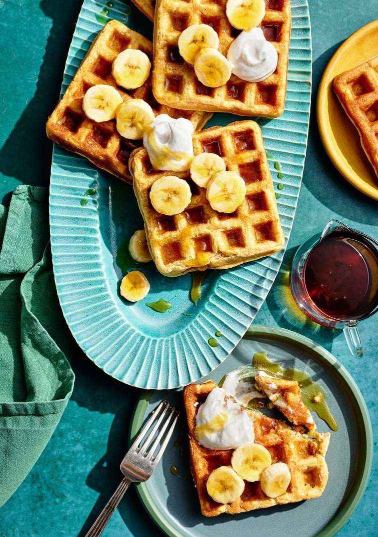 Overhead shot of waffles with banana slices and whipped cream on turquoise and blue plates, with a green napkin and syrup.
