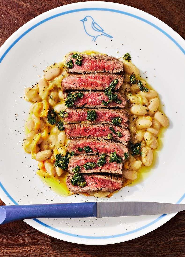 Overhead shot of sliced steak drizzled with pesto, served with beans on a white plate with blue trim and accompanied by a knife.