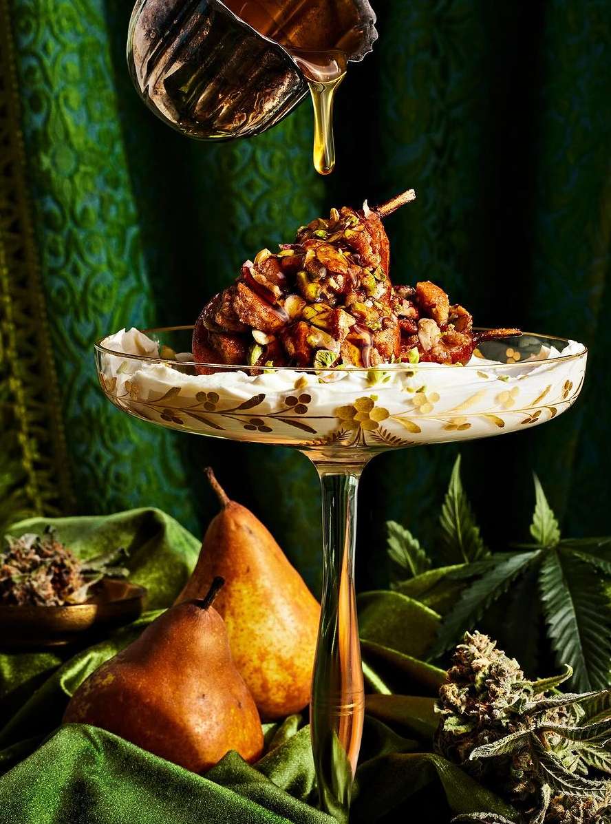Gourmet dessert presentation featuring poached pears on a stemmed glass, drizzled with honey, and garnished with green herbs against a green background.