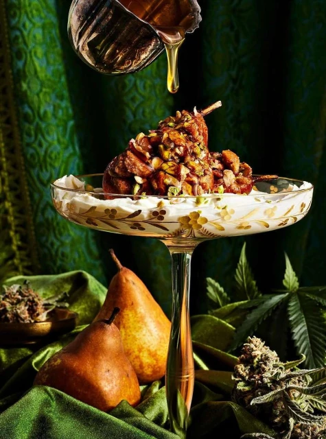 Gourmet dessert presentation featuring poached pears on a stemmed glass, drizzled with honey, and garnished with green herbs against a green background.