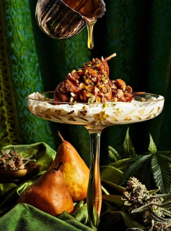 Gourmet dessert presentation featuring poached pears on a stemmed glass, drizzled with honey, and garnished with green herbs against a green background.
