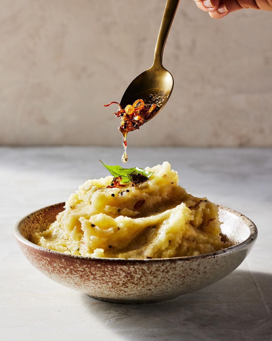 Overhead studio shot of a bowl of creamy mashed potatoes topped with a drizzle of oil, crispy fried onions, and a basil leaf.