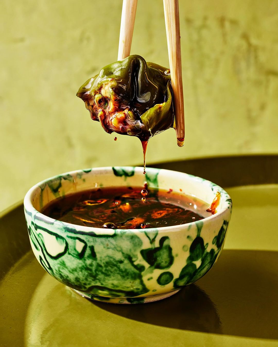 Dumpling held in chopsticks dripping sauce into bowl. Food photography showing savory appetizer and sauce against olive green background.