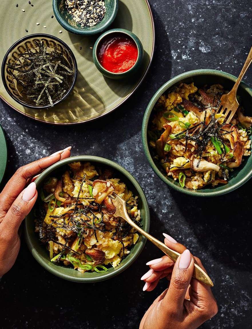Overhead shot of two bowls of Asian-style egg and vegetable rice with a dark-skinned person holding a fork in one of the bowls.