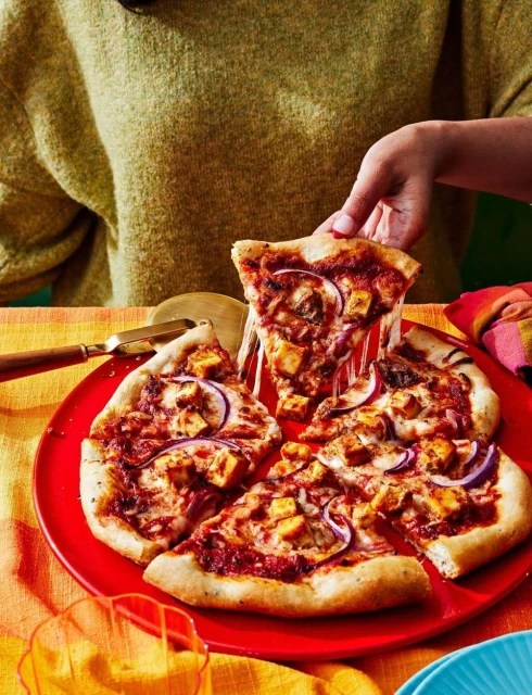 Overhead shot of a person taking a slice of homemade pizza with red onions, chicken, and cheese from a red plate on a table.