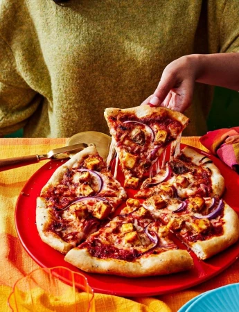 Overhead shot of a person taking a slice of homemade pizza with red onions, chicken, and cheese from a red plate on a table.
