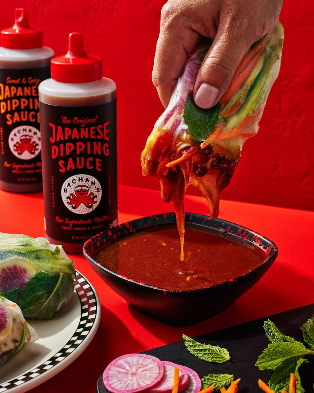 Close-up of a hand dipping a spring roll into a bowl of Japanese dipping sauce with bottles of Bachan's sauce on a red background.